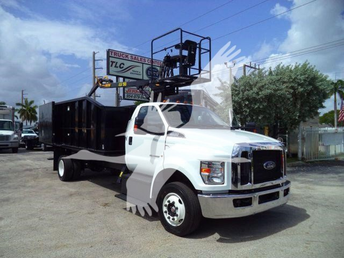 Truck with a white cab and black dump bed featuring a raised forklift-style crane on top; Ford emblem visible on grille; surrounding lot with trees and signs.