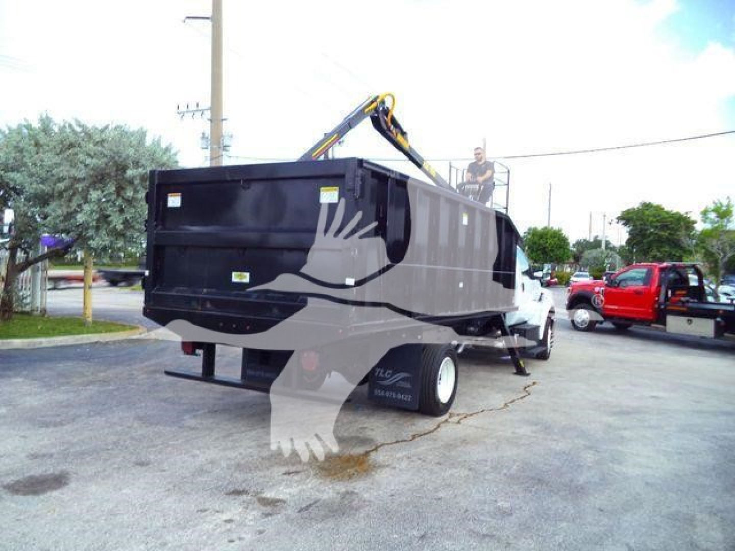 Dump truck/grapple truck in a lot showing a large black dump body with a hydraulic grapple arm on top; rear view with tailgate, side decals, and a person on the cab area.