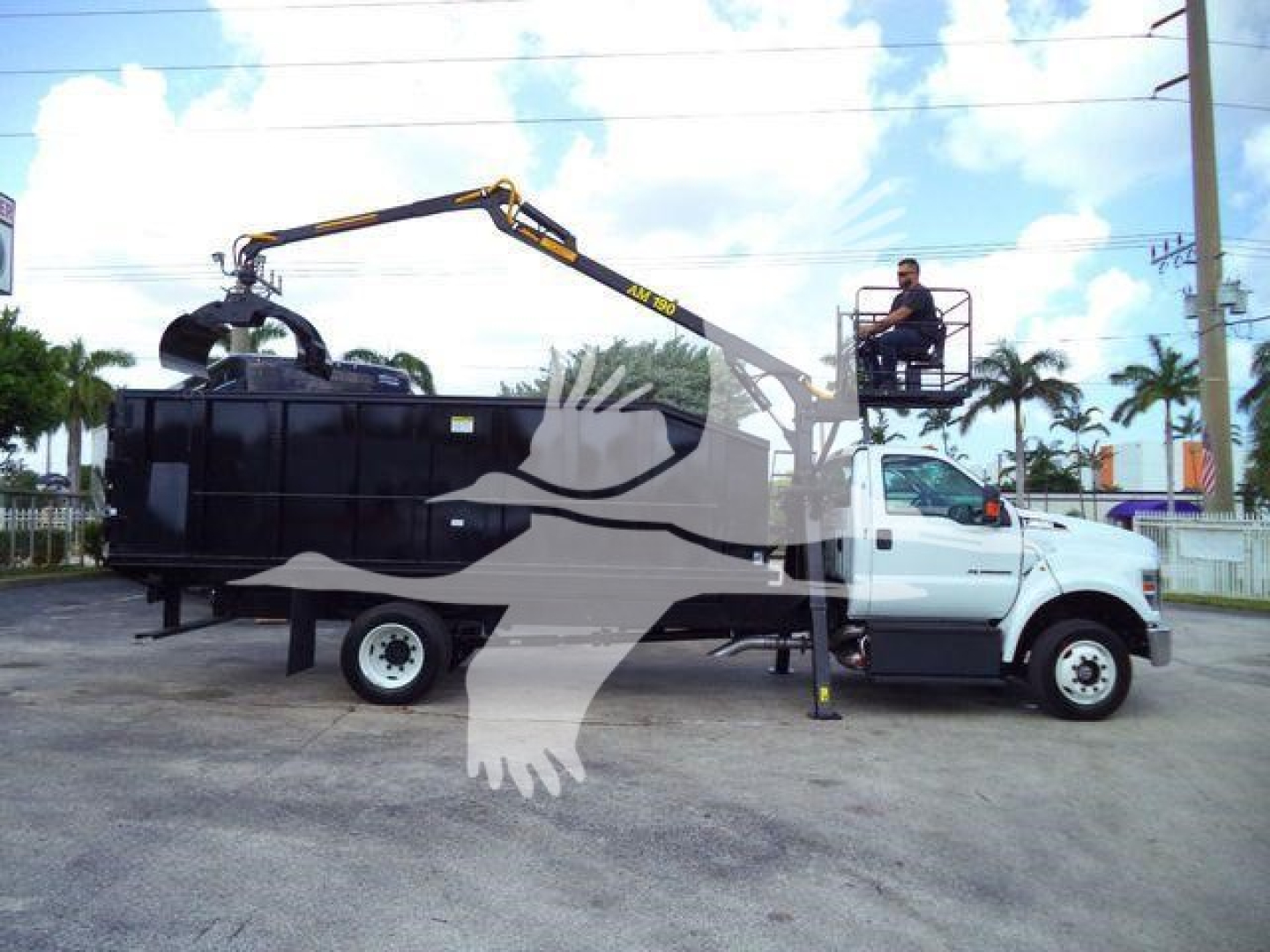Truck-mounted grapple truck with a white cab and a large black container body, hydraulic arm extended over the container, worker on a platform near the cab.