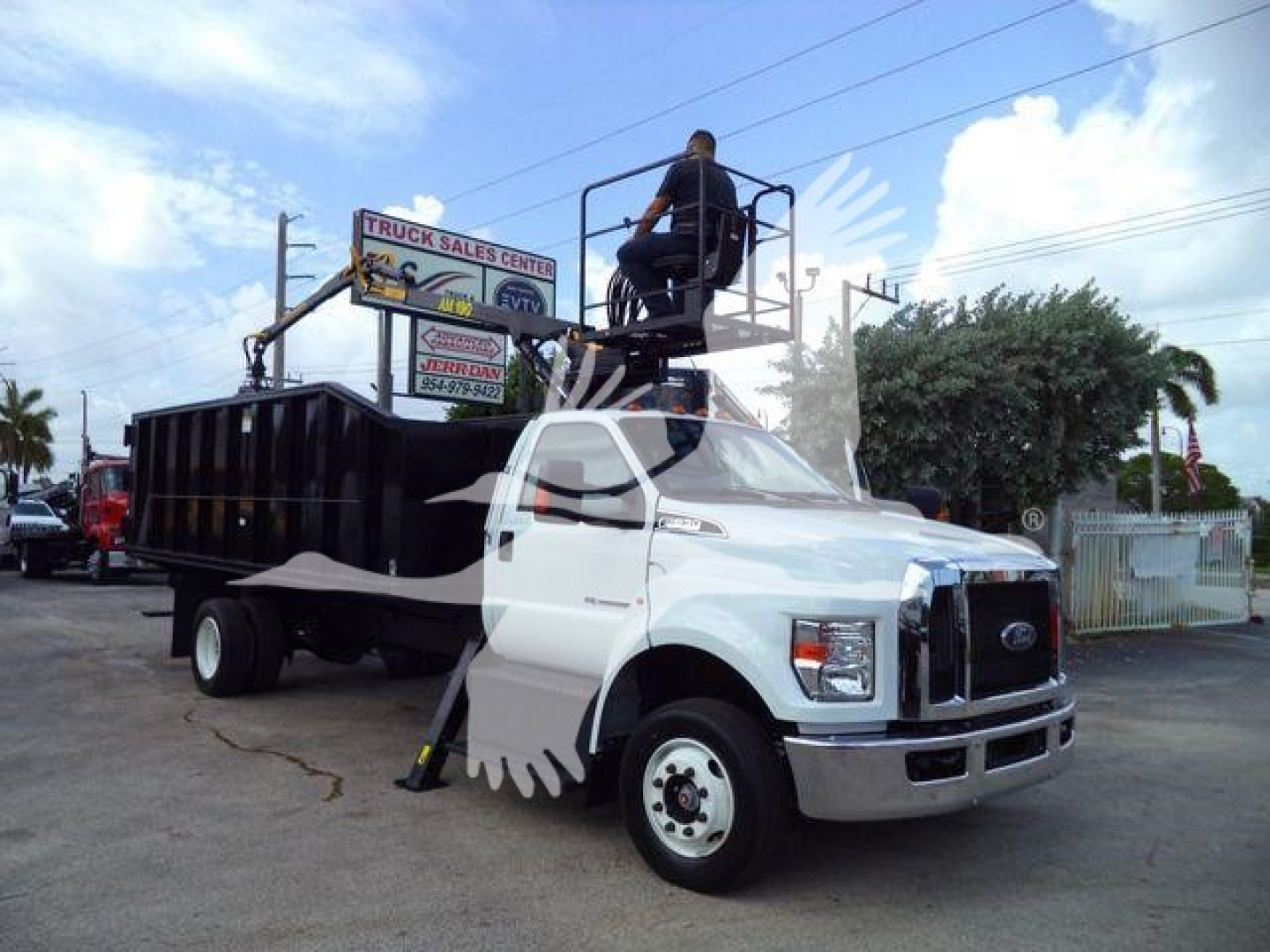 Truck with boom grapple on back and man in elevated platform; signage in background says 'TRUCK SALES CENTER' and various dealership signs
