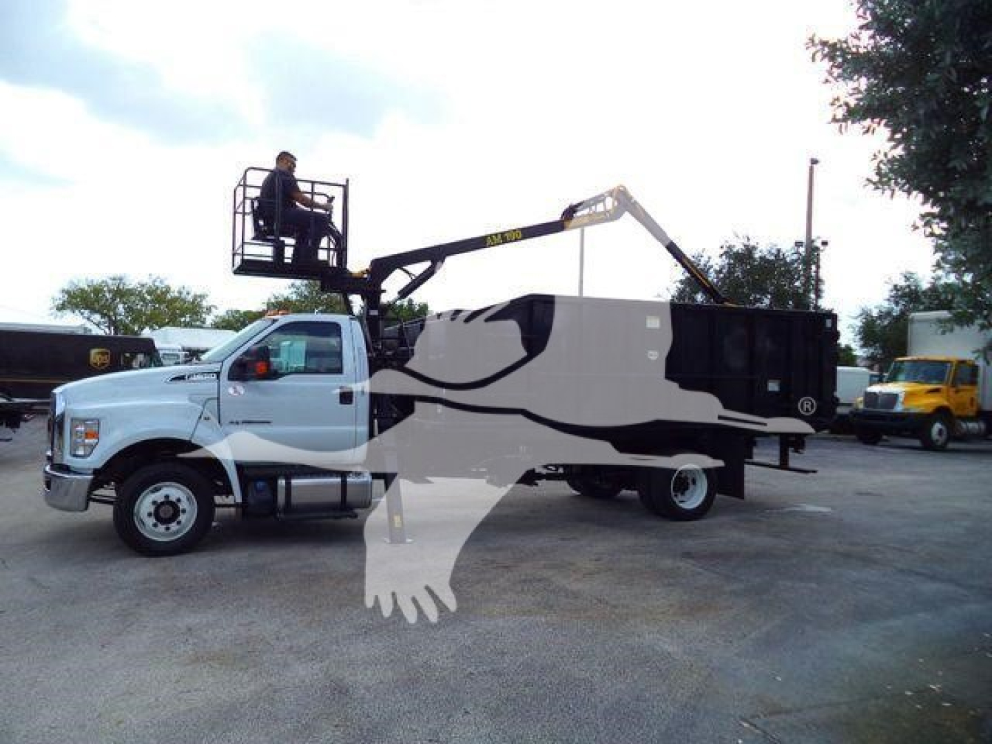 Truck-mounted grapple with white cab and black-and-gray boom in a storage lot; operator elevated in platform; rear grapple arm extended over an enclosed body.