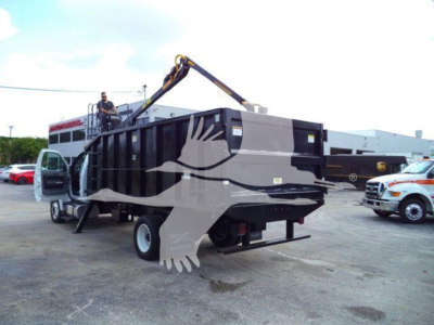 Ford F650 dump truck with grapple arm in yard, side of truck open, man on crane performing operation, black/gray body with white accents.
