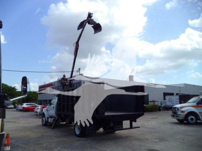 Truck-mounted grapple crane on a Ford F650 chassis with a large white bird graphic overlay on the side; equipment and dealership lot in background.