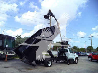 Ford F650 grapple truck with front-end bucket/attachment lifting a large metal bucket, mounted on a white truck chassis, in a dealership lot