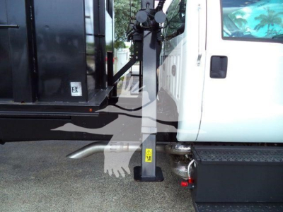 Truck side view showing a white cab and black grapple unit with yellow warning labels on stabilizer. Ground-level view with hydraulic arm attached.