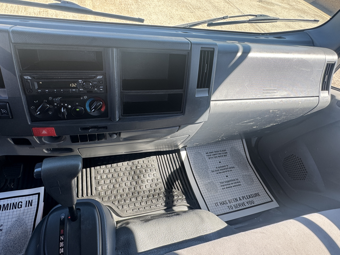 Isuzu NPR-HD interior dashboard view showing steering column, center console, climate controls, gear shifter, and empty storage slots; passenger side floor mat and dashboard details visible.