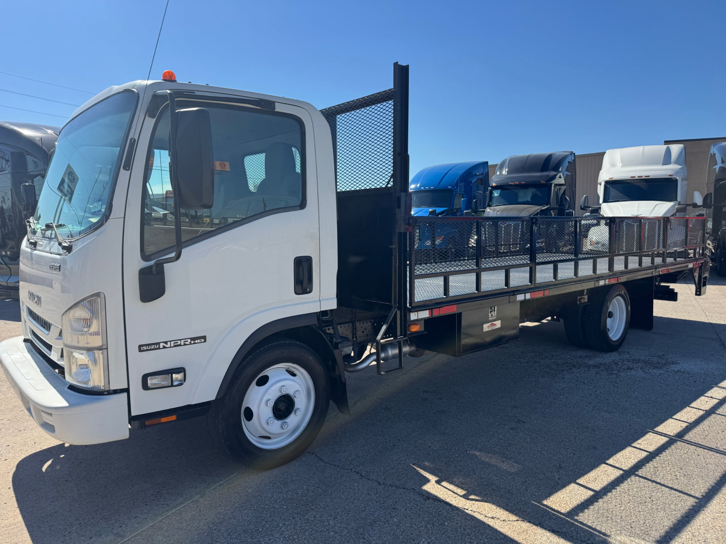 Isuzu NPR-HD flatbed truck with cab and long metal flatbed, black mesh divider behind cab, white cab, stock number plate visible on door, blue and white trucks in background.