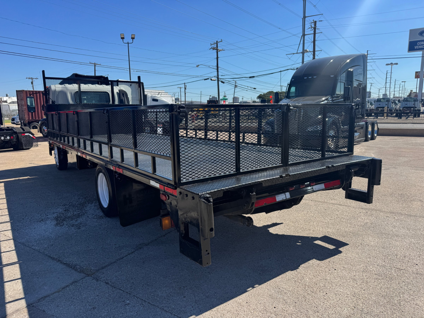 Isuzu NPR-HD flatbed truck with a black powder-coated utility gate and mesh side rails in a parking lot.