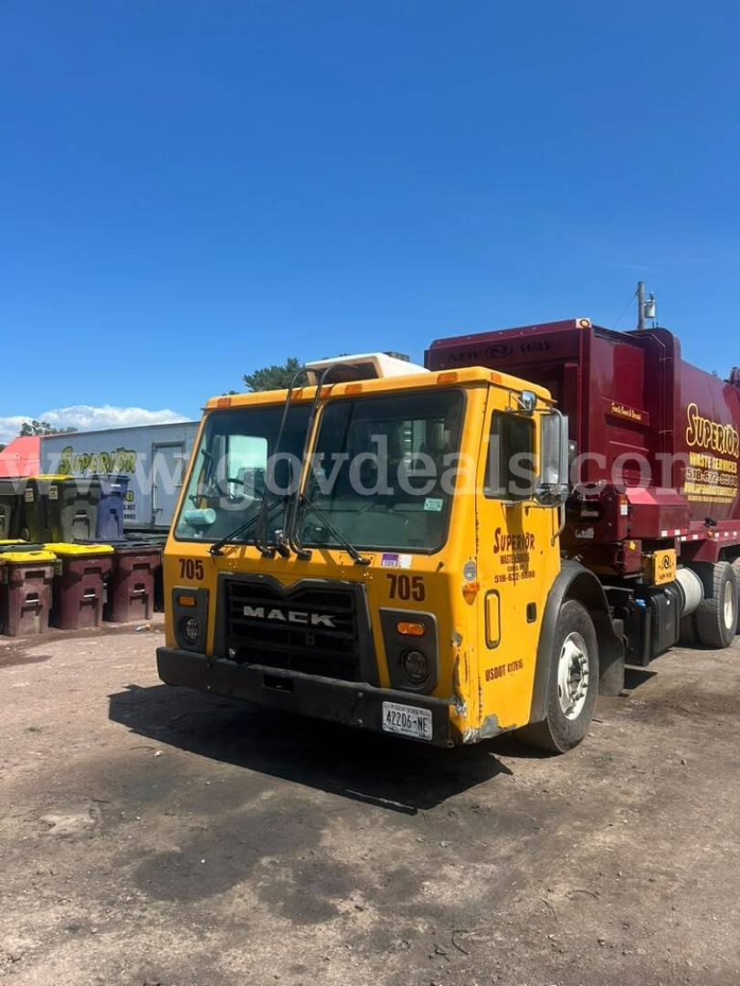 Truck front view of a yellow Mack LEU600 with a large red waste container attached.