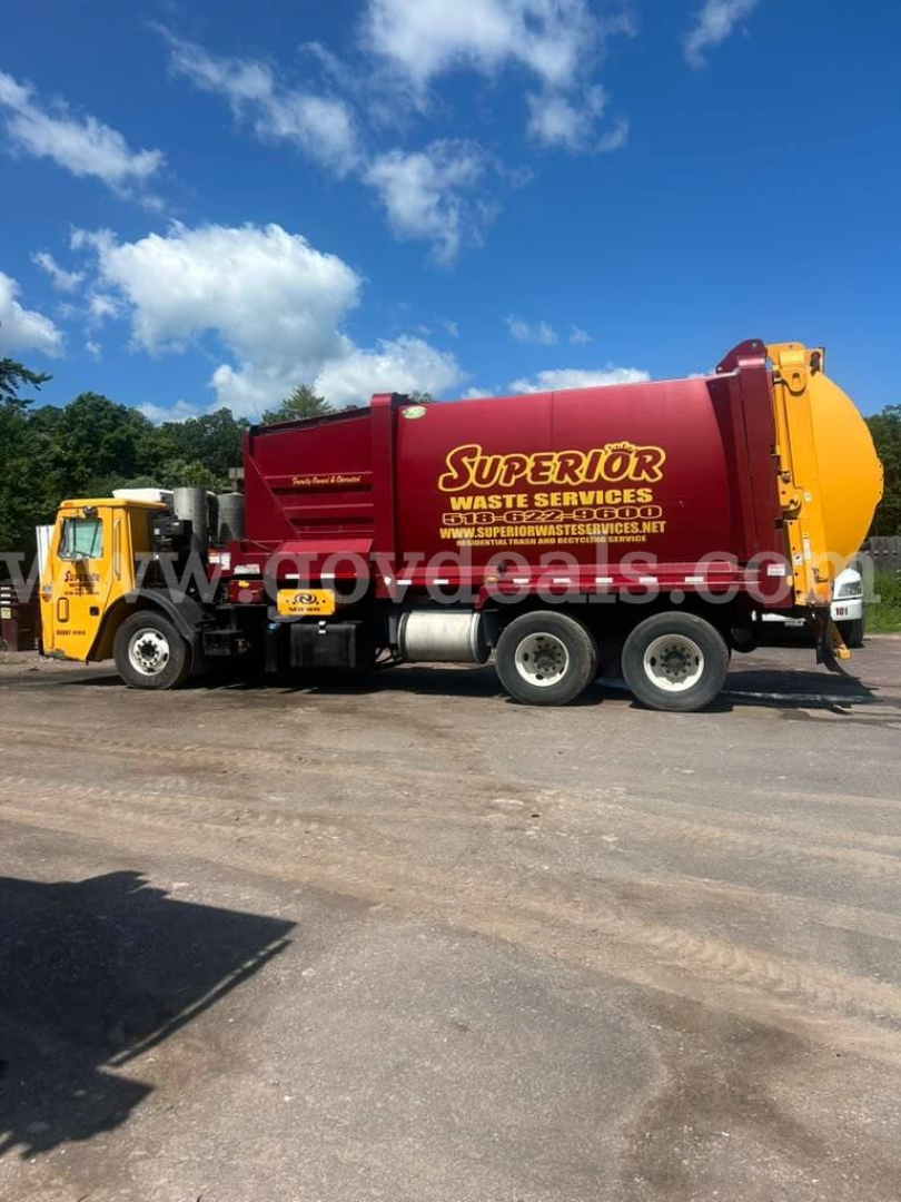 Truck side view of a yellow cab attached to a red waste liquid tanker with 'Superior' branding. Visible text includes 'Superior Waste Services' on the tanker and web address. The vehicle is a Mack LEU600 model, with a yellow cab and red/ora