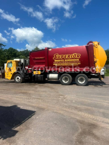 Truck side view of a yellow cab attached to a red waste liquid tanker with 'Superior' branding. Visible text includes 'Superior Waste Services' on the tanker and web address. The vehicle is a Mack LEU600 model, with a yellow cab and red/ora