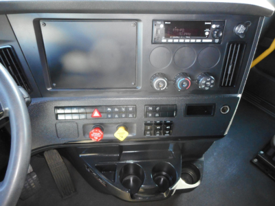 Cab interior of a Freightliner Cascadia showing dashboard with large touchscreen display, climate controls, audio head unit, ignition area, and cup holders.