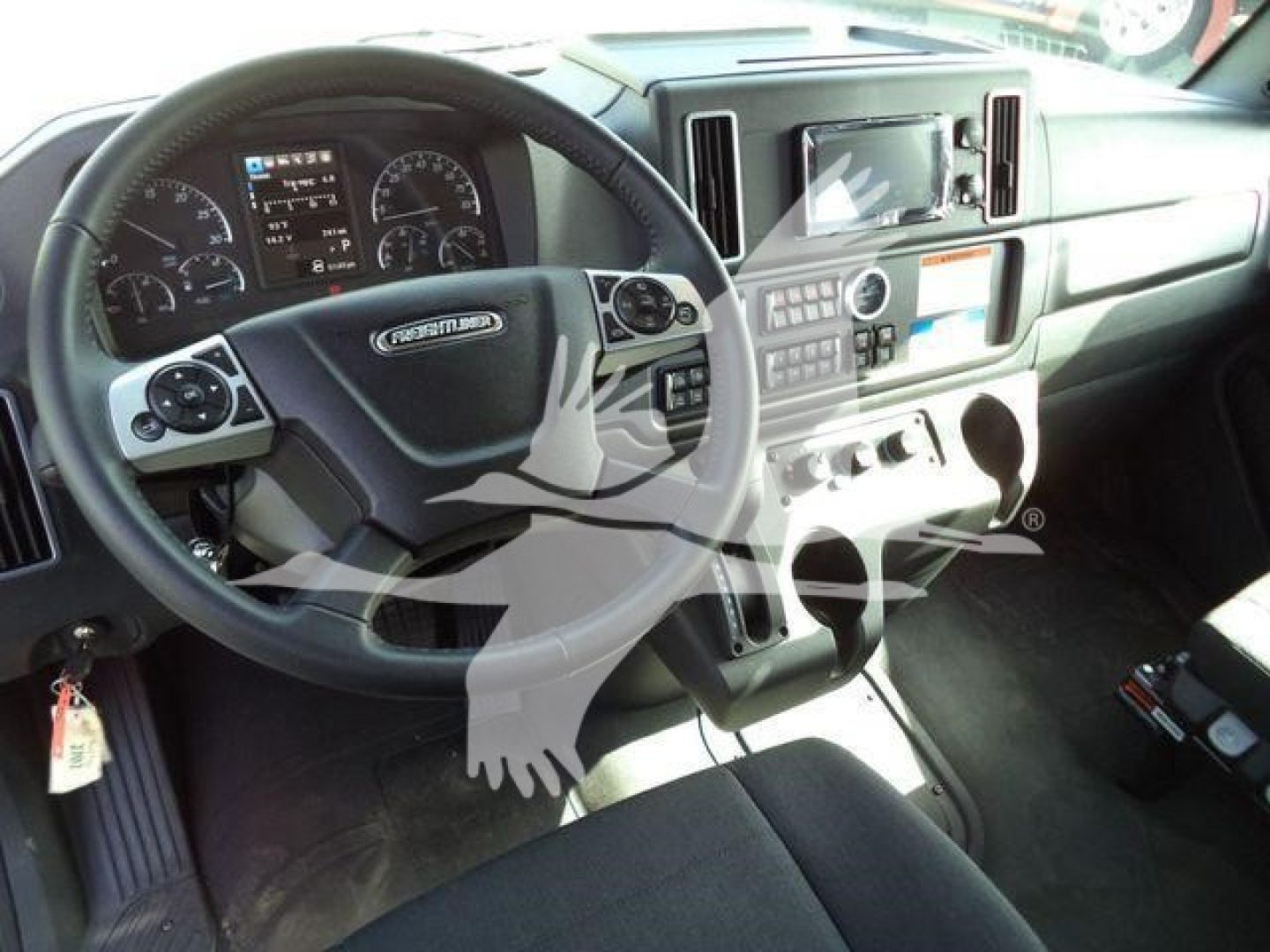 Truck cab interior showing steering wheel with Freightliner badge, dashboard, central console with touchscreen, gear controls, and cup holders.