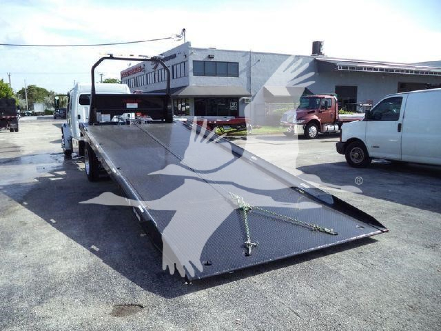 Freightliner rollback truck bed in a dealership lot, with a long flatbed and ramp in view; surrounding white and brown service vans and a building in the background.