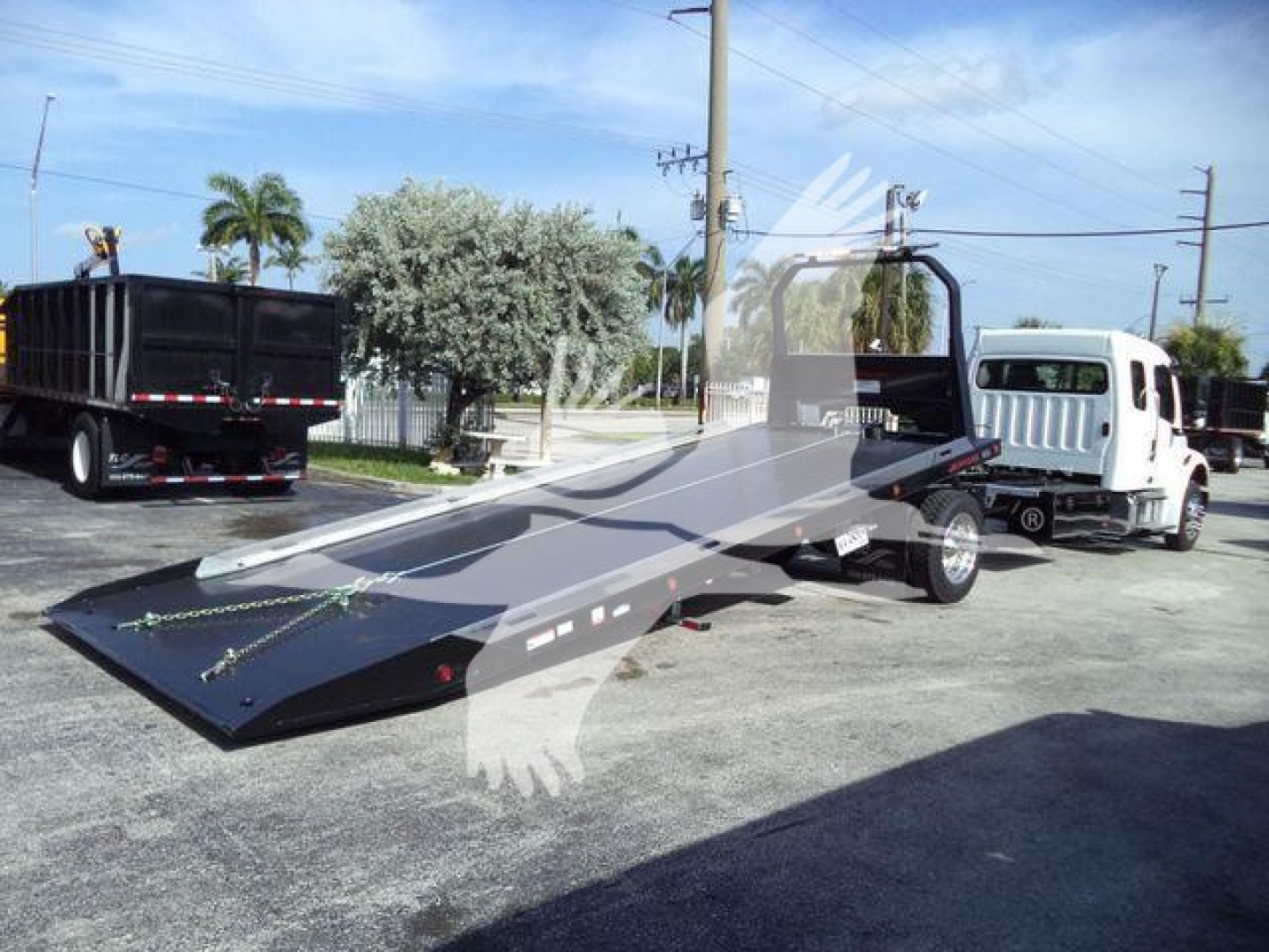 Freightliner Business Class M2 106 rollback tow truck with a tilted flatbed in a dealership lot; white cab, black drop-bed, other trucks in background.