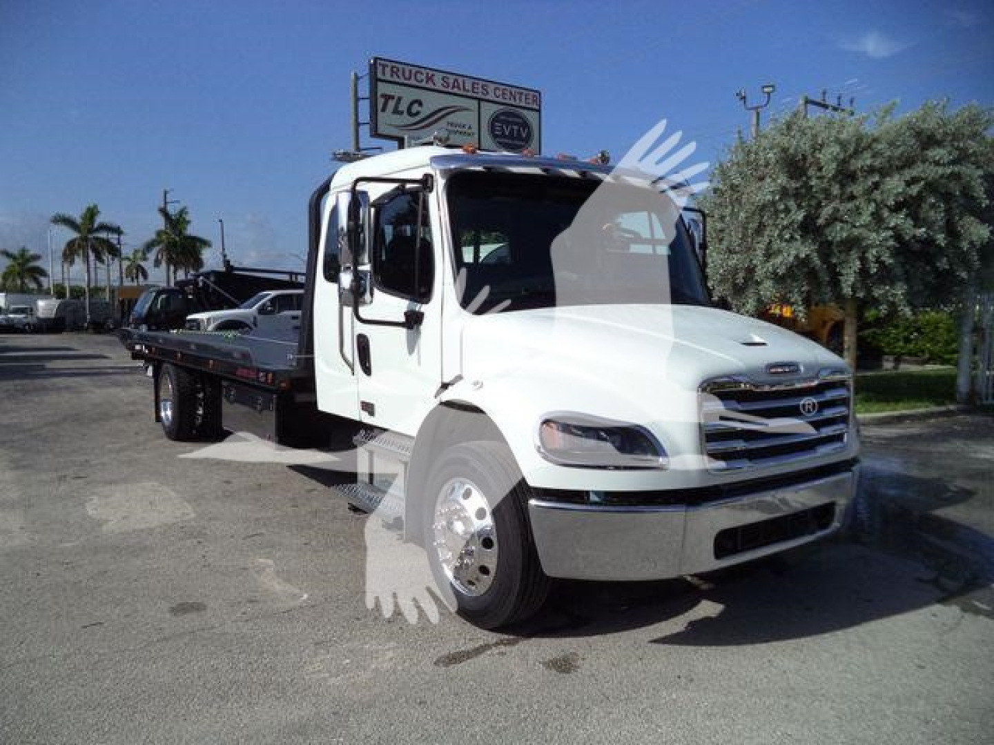 Freightliner business class M2-106 rollback tow truck in white with flatbed, front view, logo on grille, dealership lot in background.
