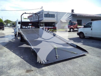 Freightliner rollback truck bed in a dealership lot, with a long flatbed and ramp in view; surrounding white and brown service vans and a building in the background.