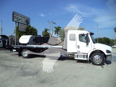 Freightliner rolling heavy-duty rollback tow truck side view showing cab and flatbed with wheel area and sign in background.