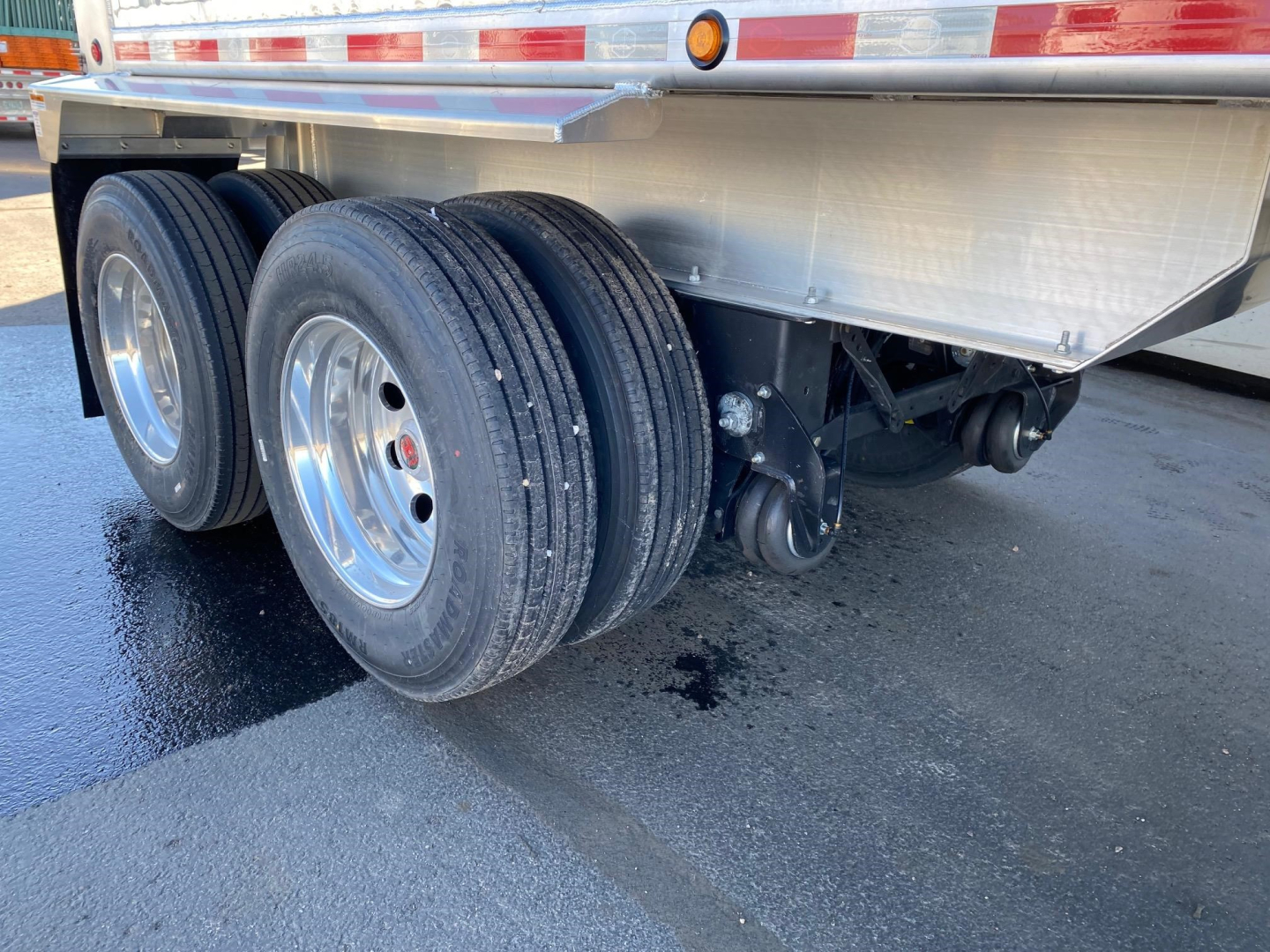 Trailer axle group showing dual rear axles with three tires and chrome rims under a dump trailer with reflective side rails and undercarriage components visible.