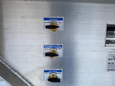 Trailer equipment panel showing tailgate, suspension dump, and lift axle controls with labels and gold toggle levers on a metallic surface.