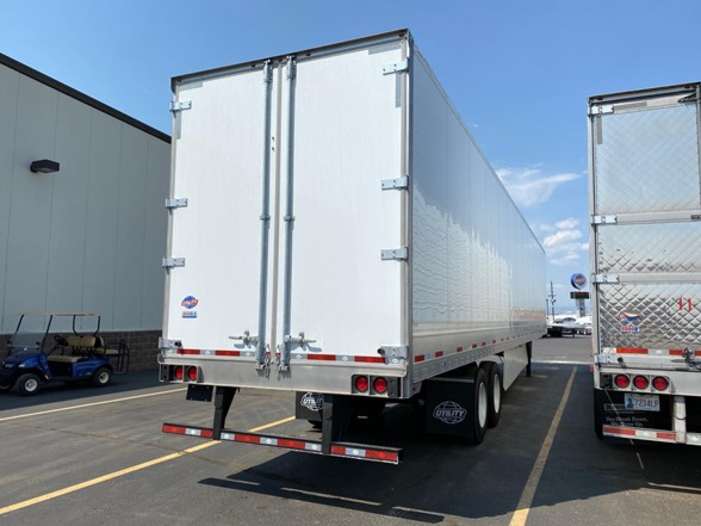 Trailer rear view showing two white cargo doors, taillights, and dual rear wheels. Flatbed with reflective tape and bumper undercarriage visible.