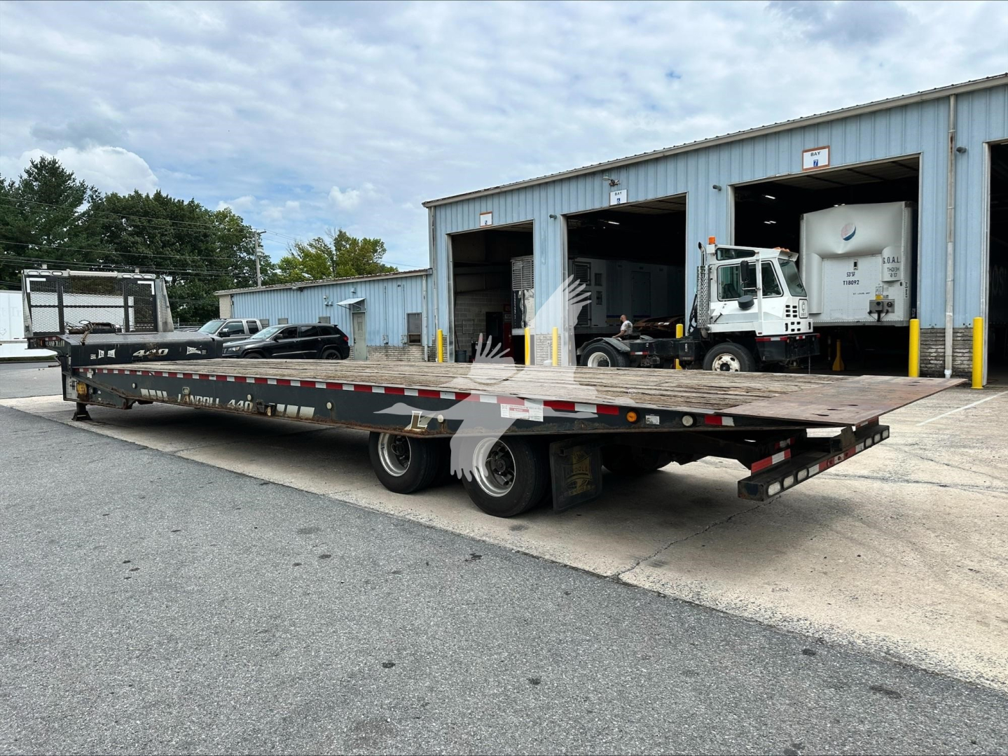 Landoll 440B trailer in a yard showing a long flatbed with dual rear axles and wheel ramps, warehouse buildings in the background.