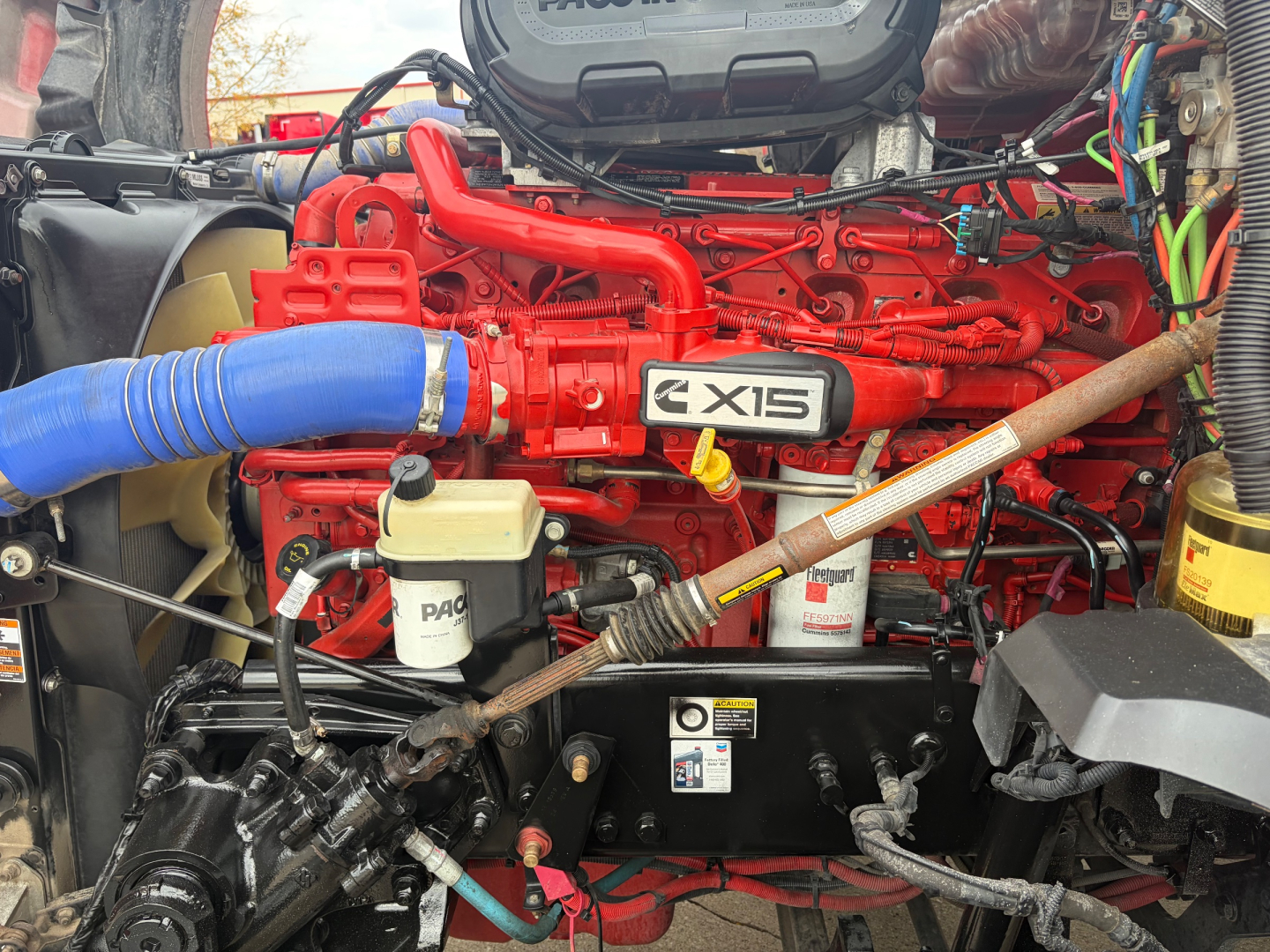 Kenworth T680 engine bay showing ISX12G red engine with blue intake hose and various reservoirs and wiring; branding visible on engine cover.