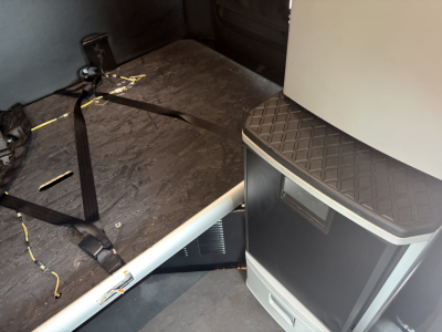 Interior of a 2023 Kenworth T680 cab showing a sleeper area with a seat or platform and a step; visible firewall and plastic trim. No engine or labels clearly legible in this shot.