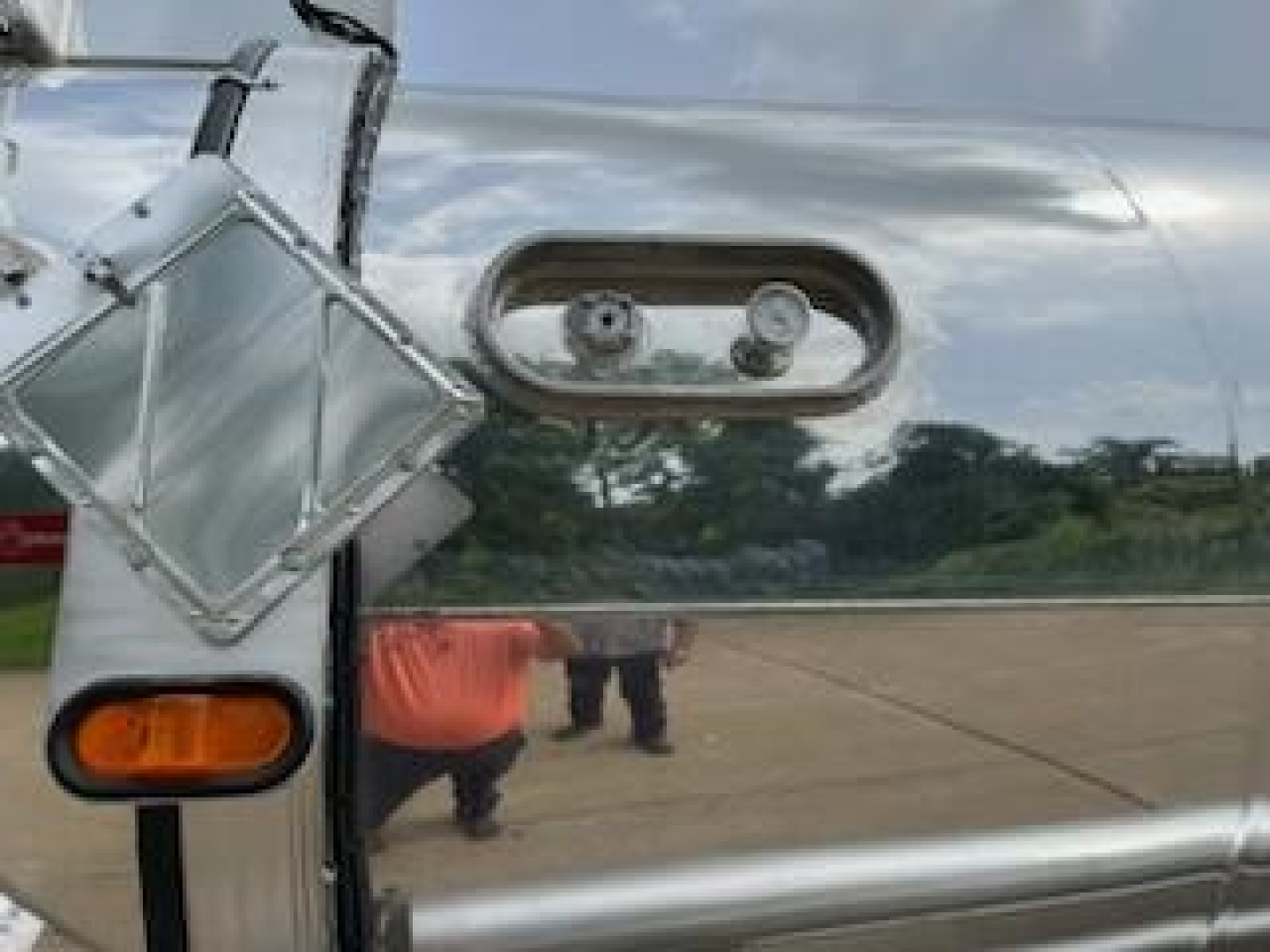 Trailer fuel or equipment hatch with two circular access ports on polished metal surface. Reflection shows person in orange shirt and outdoor tarmac.