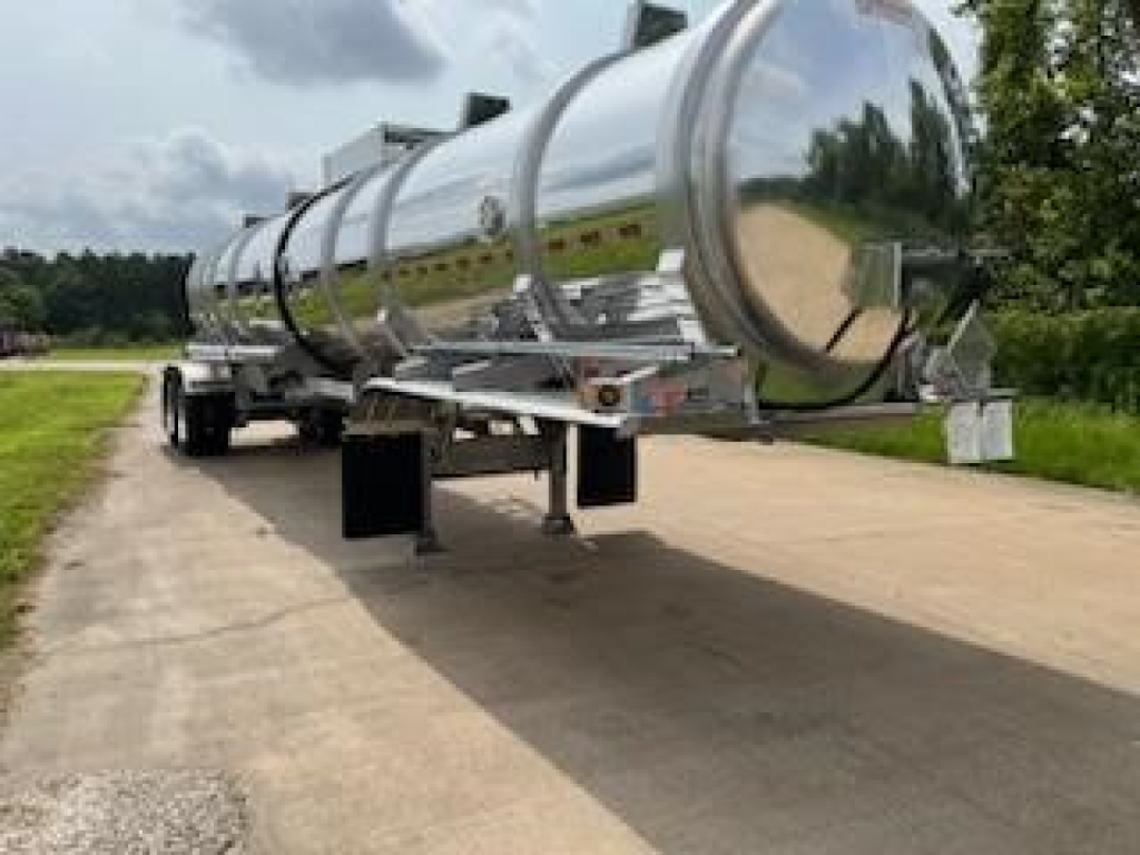 A shiny stainless steel tanker trailer on a paved road, two-axle cylindrical tank with supporting legs and front hitch visible.