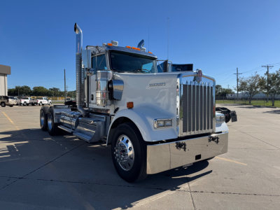 Kenworth W900L conventional sleeper truck front-left view showing large chrome grille, vertical exhaust stacks, dual rear axles, and chrome bumper with clear blue sky.