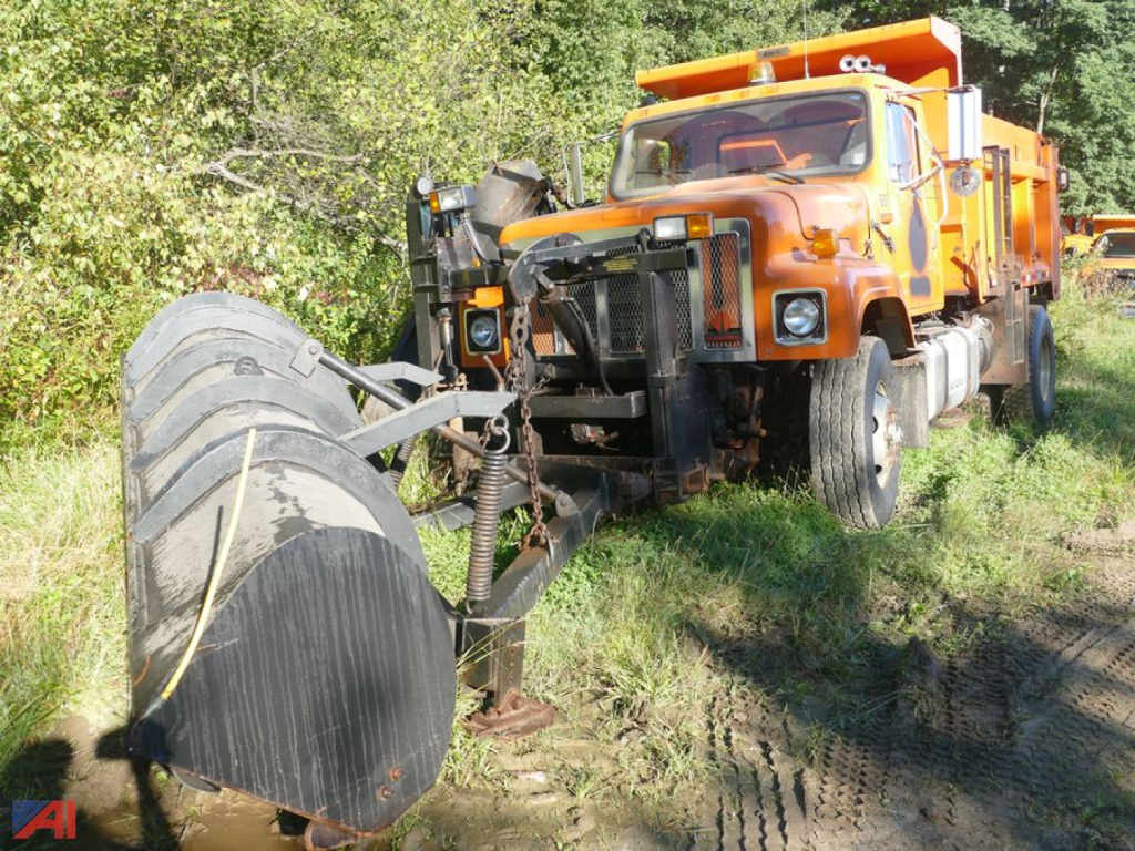 Front view of an orange 2002 International dump truck with a large front plow/scoop and mounted attachment in the foreground; headlights, grille, and cab visible.