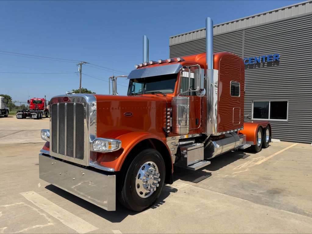Peterbilt 389 conventional sleeper truck in orange, front left view, chrome grille, dual exhaust stacks, large polished bumper, daytime parking lot.