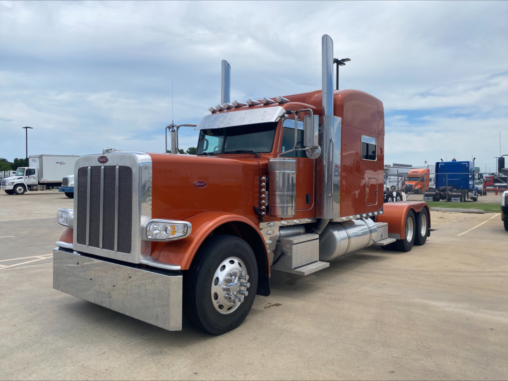 Peterbilt 389 tractor with orange cab and sleeper, chrome exhaust stacks, large front grille, and dual rear axles, parked in a lot.
