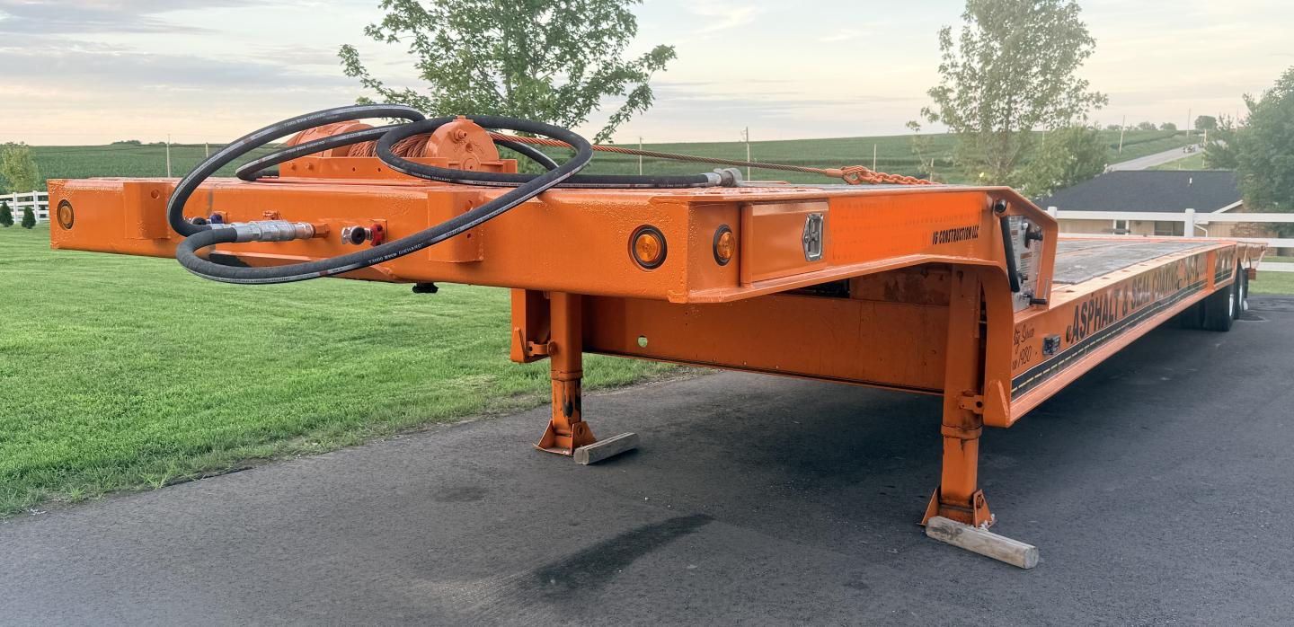 Traveling tail assembly of a Landoll lowboy trailer in orange, with a circular control wheel and hydraulic hoses visible on the front, side rails and landing legs in view.