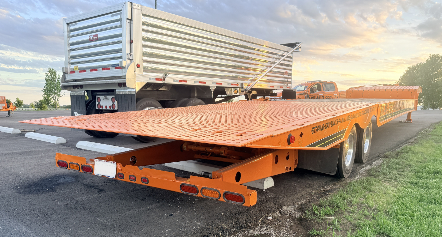 Traveling tail trailer with orange lowboy deck and ramp catwalks, rear view showing hydraulic mechanism and taillights.