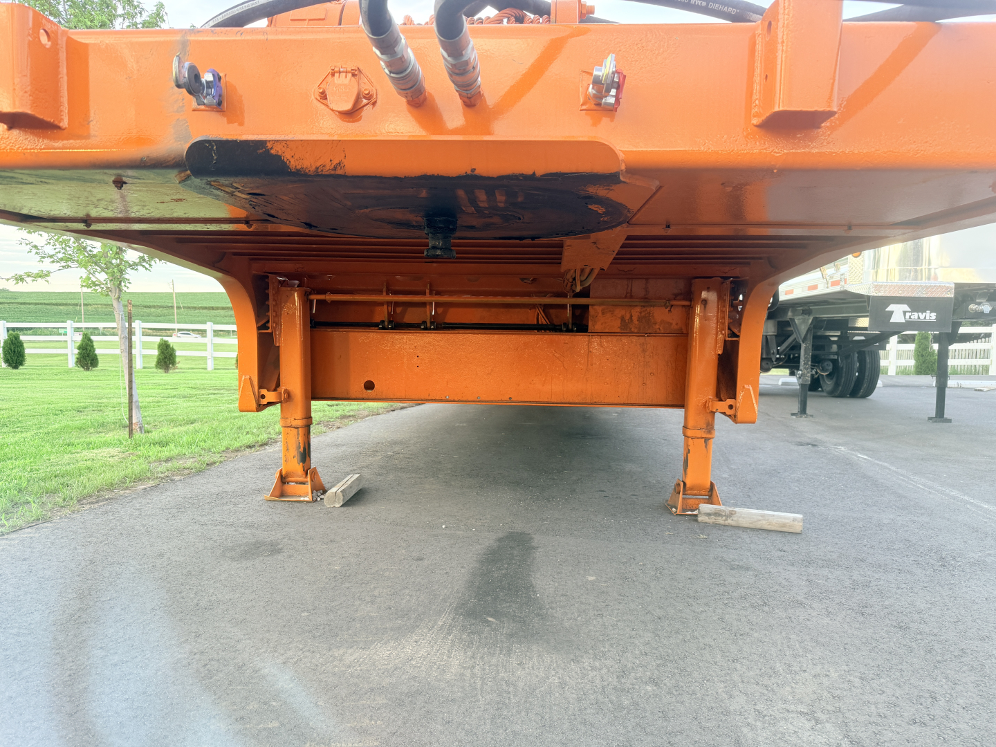 Rear view of an orange Landoll 930-51-15 traveling tail trailer on a paved lot, showing landing gear and underside with hydraulic hoses and frame.
