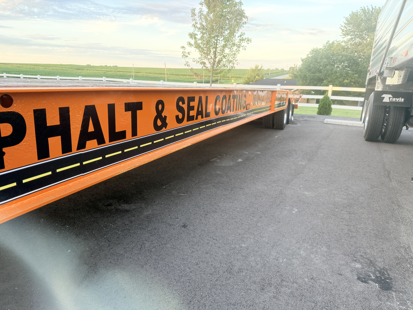 Long orange Landoll lowboy trailer with black text reading PHALT & SEAL COATING and a truck close by.