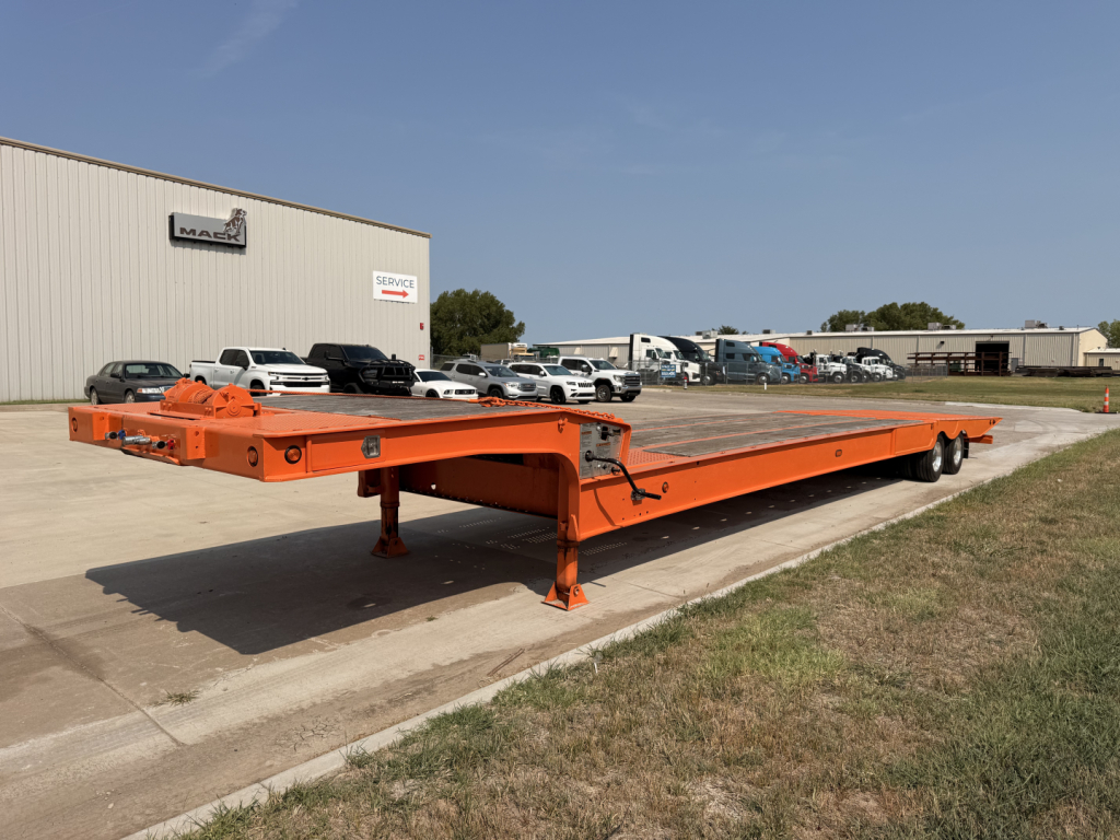 Landoll 930-51-15 traveling tail lowboy trailer in orange, long deck with landing legs and dual axles; shows flatbed surface and equipment yard background.