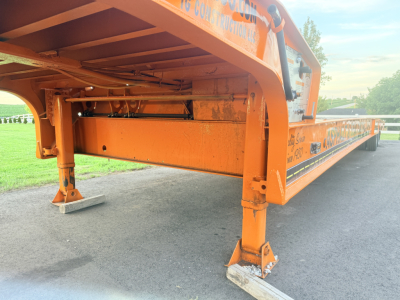 Traveling tail lowboy trailer with orange frame and adjustable hydraulic legs, side labeling visible.