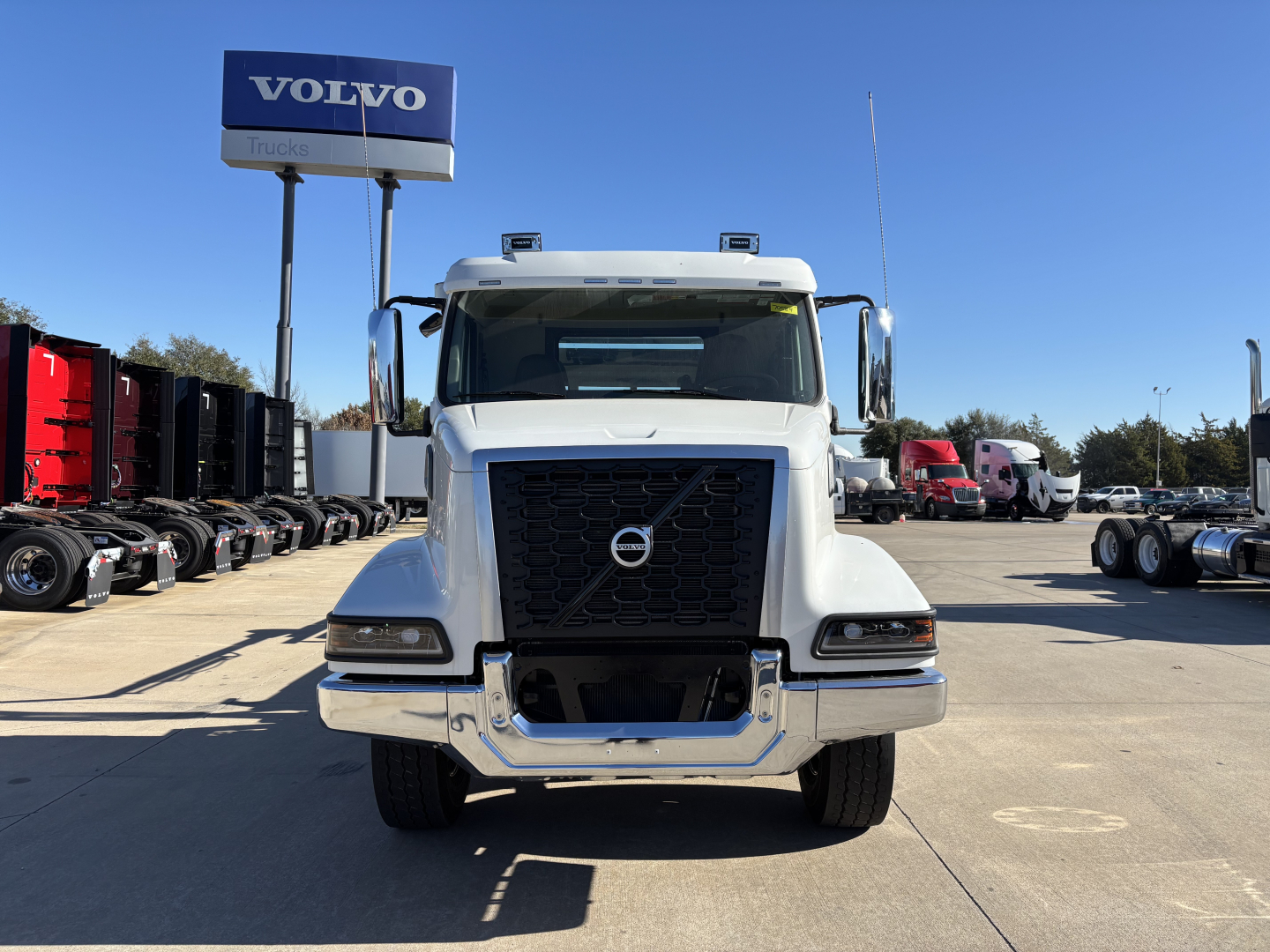 Front view of a white 2026 Volvo VHD64B300 truck in a dealership lot, Volvo sign in background, prominent black grille with Volvo emblem, large bumper, dual mirrors.