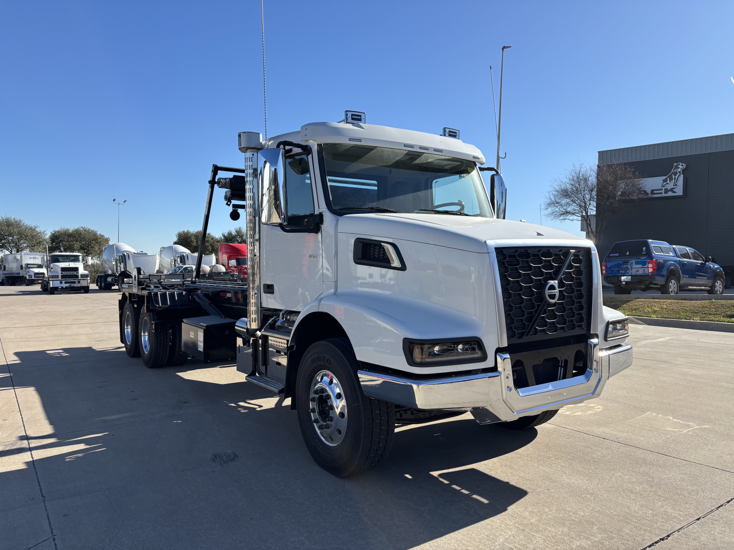 Volvo VHD64B300 rolloff truck in a dealership lot with white cab and flatbed, blue sky, VIN 4V5KC9DH1TN710407 and stock number 70584 visible.