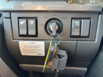 Vehicle ignition area of a Volvo VHD64B300 engine bay control panel with key in ignition and two toggle switches on left and right, warning label below.