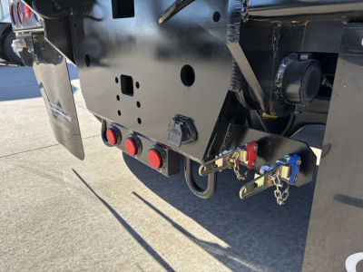 Volvo VHD64B300 roll-off truck rear chassis showing hitch, hydraulic lines, and safety latches with red/blue components and chain attachments.