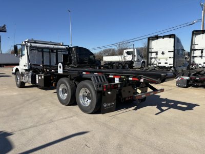 Volvo VHD64B300 rolloff truck chassis side and rear with hydraulic bed and dual rear axles, white cab, black flatbed, visible tires and taillights.
