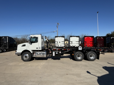 Volvo VHD64B300 rolloff truck side view with white cab and long flatbed, showing VIN 4V5KC9DH1TN710407 and stock area in background.