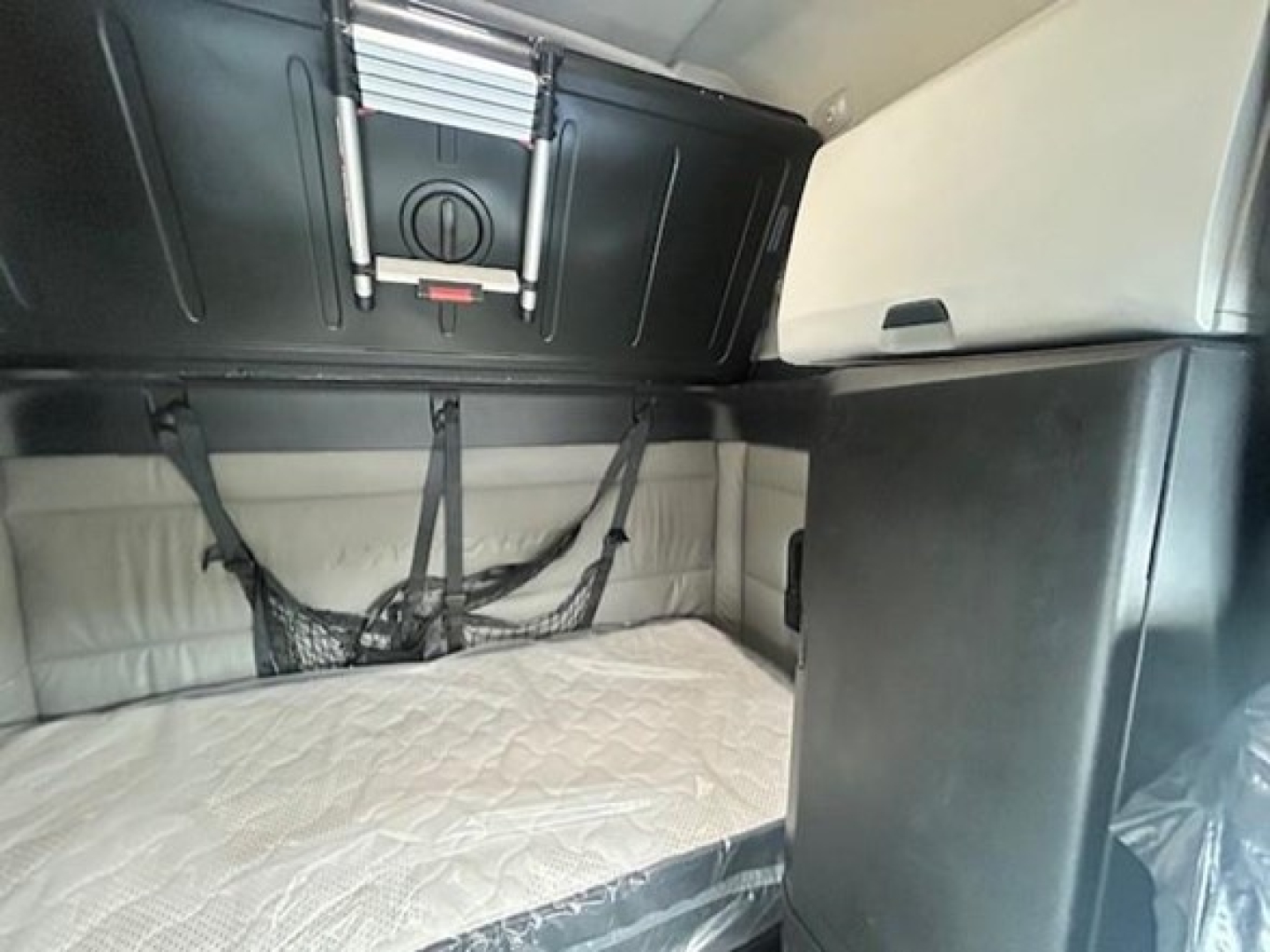 Truck cab interior showing bunk area with beige mattress and overhead storage cabinet.