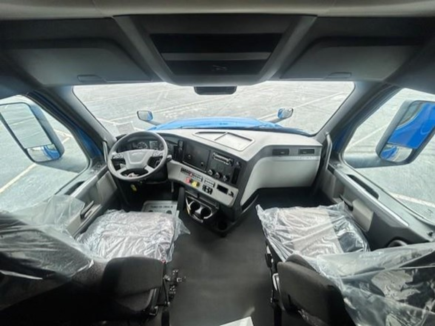 Freightliner Cascadia cockpit view showing steering wheel, dashboard with control panels, and seats wrapped in plastic.