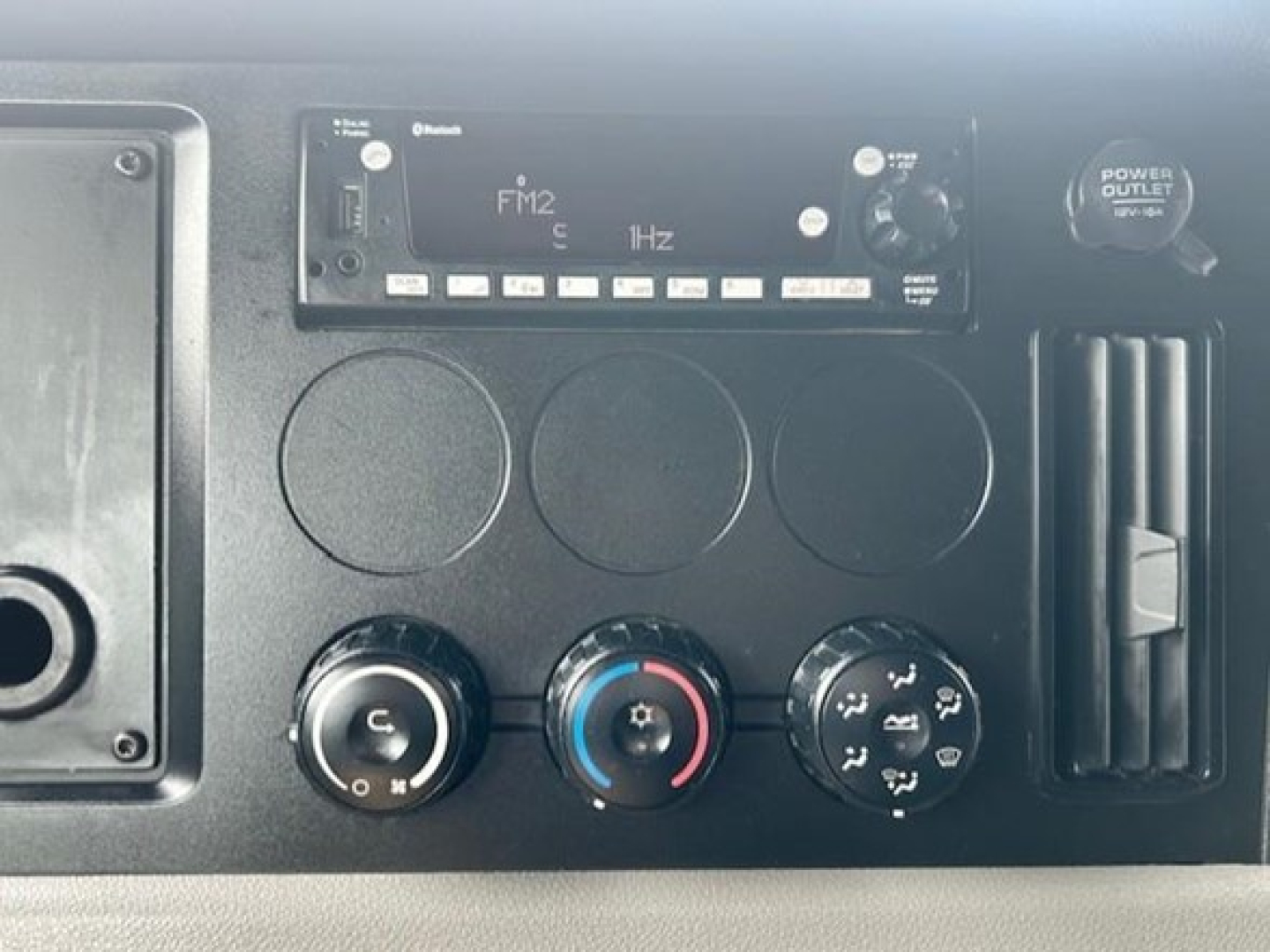 Dashboard panel of a Freightliner Cascadia showing climate controls, radio/FM2 tuner, and power outlet in a sleeper truck interior.