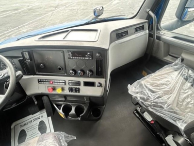 Dashboard view of 2022 Freightliner Cascadia 126 sleeper truck cab showing steering wheel, central control panel, and driver seat with plastic cover.